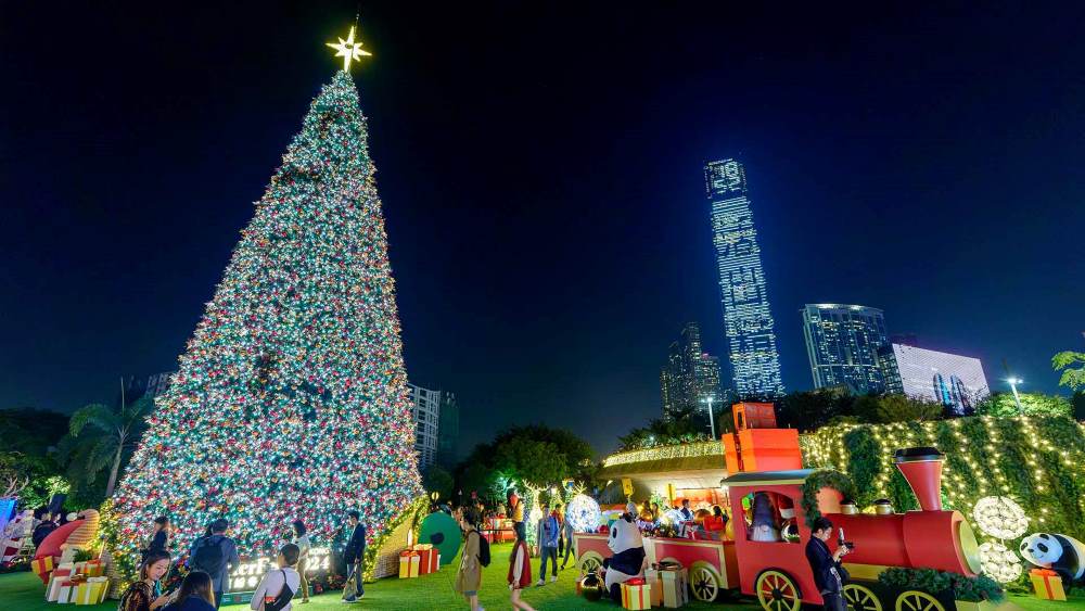 tall christmas tree with colorful lights and red train beside it with people around