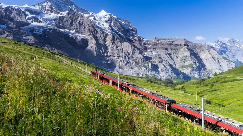 red train in the middle of a green hill in front of a mountain- Best road trips in Europe